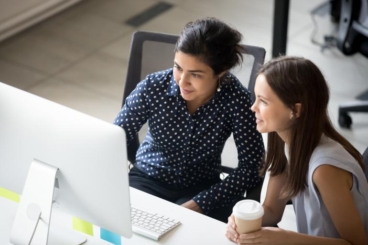 Two women collaborating at a computer desk, one holding a coffee cup and the other pointing at the screen.