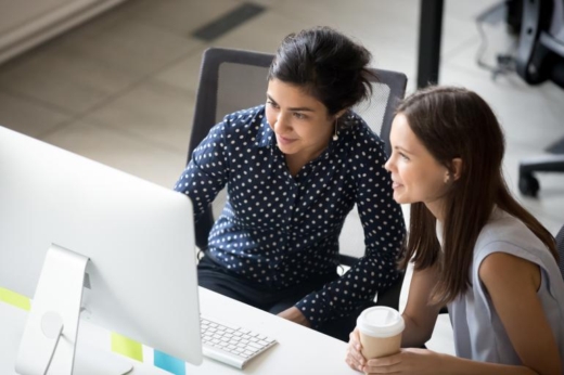 Two women collaborating at a computer desk, one holding a coffee cup and the other pointing at the screen.