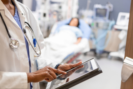 Healthcare professional in a white coat using a tablet while a patient rests in a hospital bed.