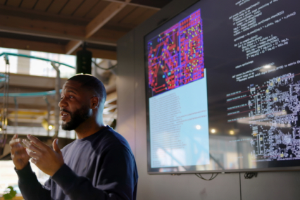 A man gesturing while presenting data and code on a large screen in a modern workspace.