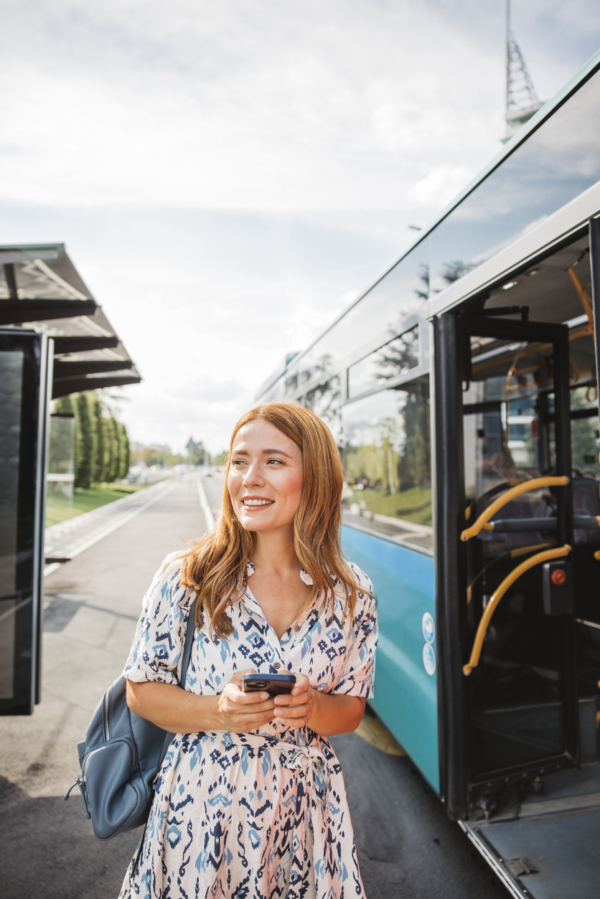 A woman in a patterned dress holding a smartphone stands next to an open bus at a station.