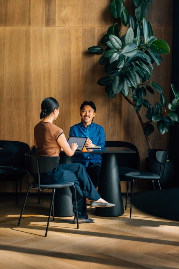 Two individuals seated at a table in a modern interior with a large plant in the background.