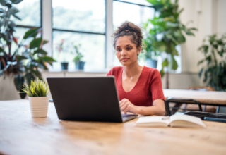 A woman sitting at a desk using a laptop, with a notebook and a plant nearby.