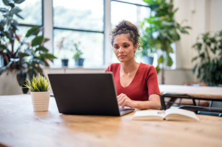 A woman sitting at a desk using a laptop, with a notebook and a plant nearby.
