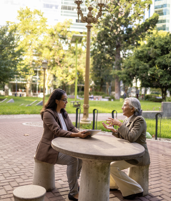 Two women sitting at a round concrete table in a park, engaged in conversation.