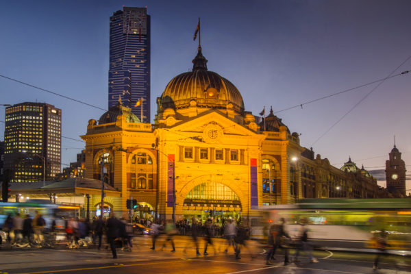 Flinders Street Station in Melbourne illuminated at dusk with people walking in front.