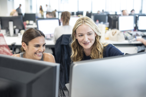 Two women smiling while working together at a computer in an office environment.