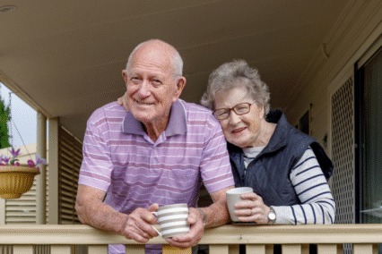 An elderly couple smiling while holding cups on a porch railing.