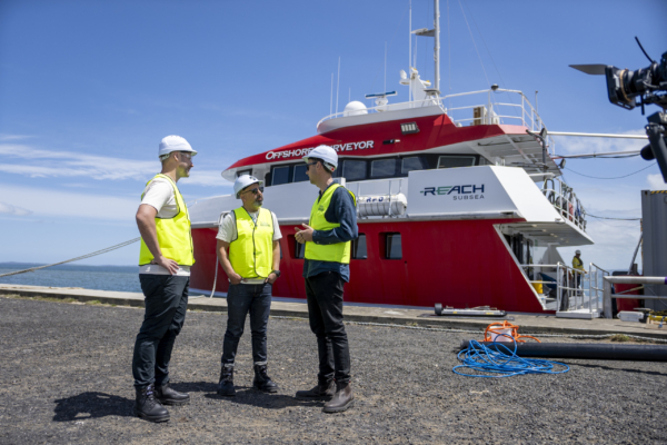 Three individuals wearing safety vests and helmets discussing near an offshore vessel named 'Offshore Surveyor'.