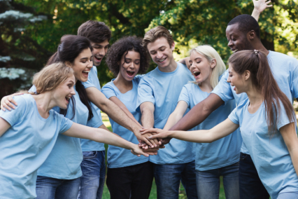 A group of young adults wearing light blue t-shirts joining hands in a circle, smiling and celebrating.