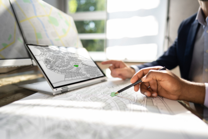 A person pointing at a digital map on a tablet while another person points at a paper map during an urban planning discussion.