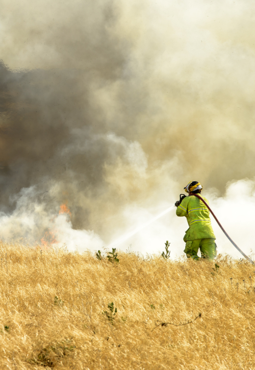 Firefighter in protective gear using a hose to extinguish a grass fire surrounded by smoke.
