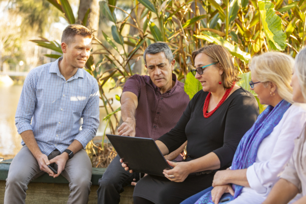 A group of four individuals engaged in a discussion while using a laptop outdoors.