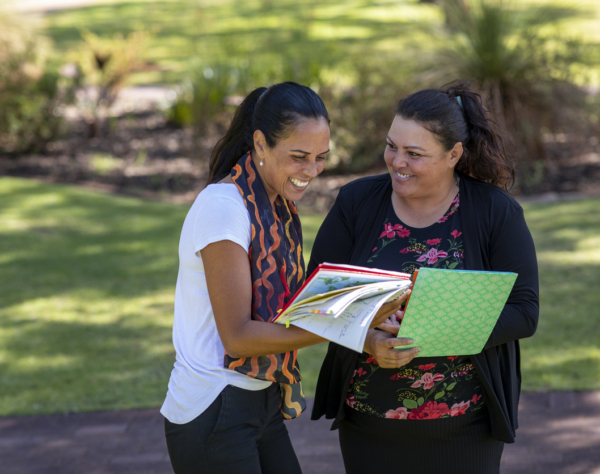 Two women smiling and reviewing documents while standing outdoors in a park-like setting.