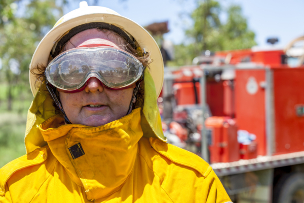 A firefighter wearing a yellow protective suit and goggles, with a fire truck in the background.
