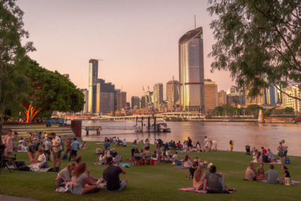 People gathered on grass in a city park near a river with skyscrapers in the background during sunset.