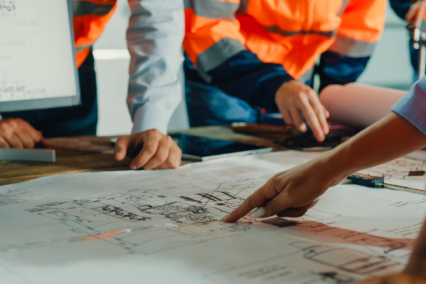 Individuals in safety vests examining construction plans on a table with a computer in the background.