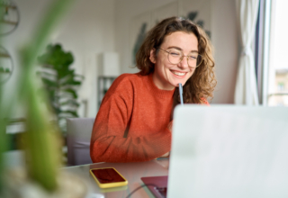 A woman with curly hair and glasses smiling while using a laptop at a desk.