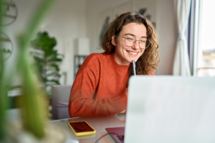 A woman with curly hair and glasses smiling while using a laptop at a desk.