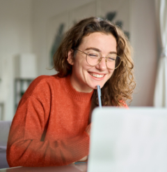 A woman with curly hair wearing glasses, smiling while sitting in front of a laptop.