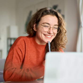 A woman with curly hair wearing glasses, smiling while sitting in front of a laptop.
