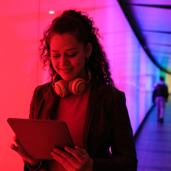 A woman with curly hair and headphones looking at a tablet in a brightly lit corridor.