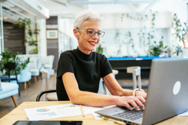 Senior woman with short gray hair typing on a laptop at a wooden table with papers and a pencil.