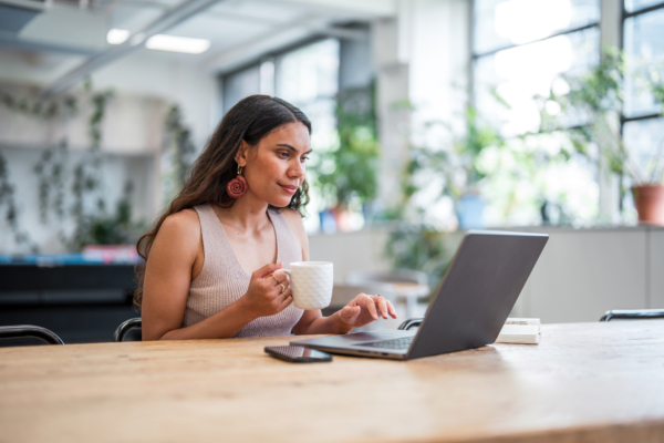 Woman sitting at a table, using a laptop while holding a coffee cup.