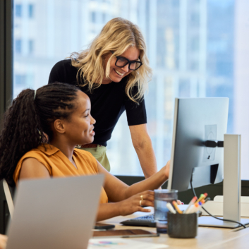 Two women engaged in a discussion while working at a desk with a computer in an office setting.