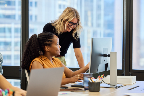 Two women engaged in a discussion while working at a desk with a computer in an office setting.