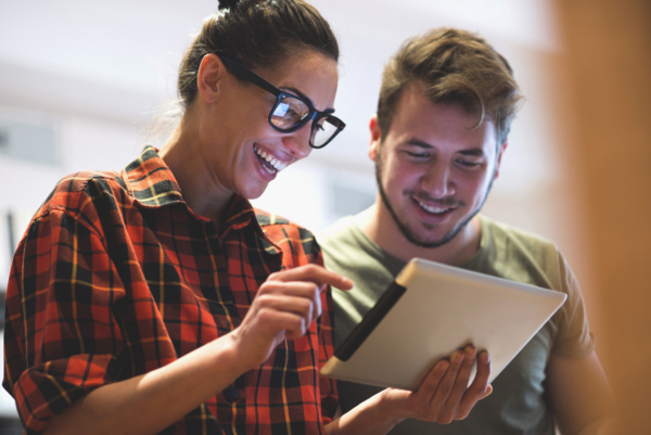 A woman in glasses and a man smiling while looking at a tablet together.