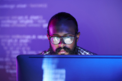 A man with glasses focused on a computer screen, with code visible in the background.