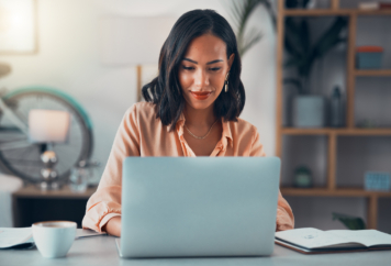A woman with shoulder-length hair working on a laptop at a desk in a modern office setting.